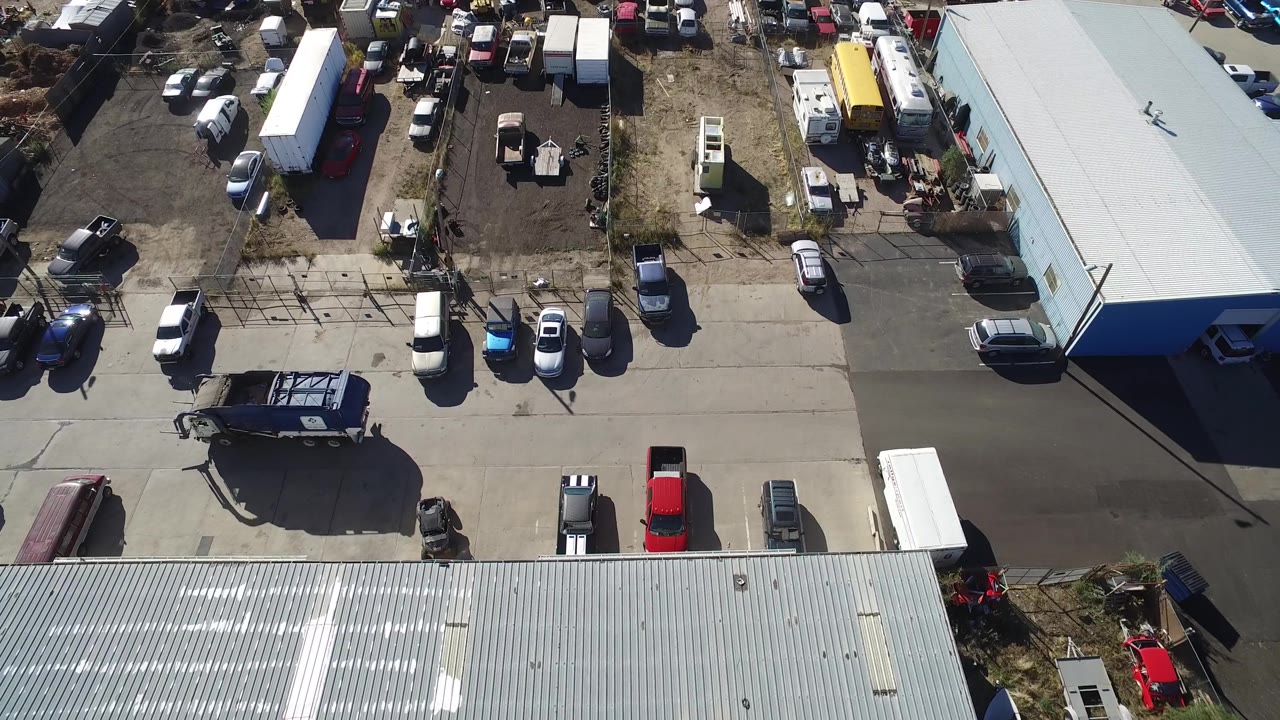 Drone-style overhead frame in Hidden Lake, Colorado with a gray roof in the foreground, parked vehicles near a fence, trailers, equipment piles, and no visible people.