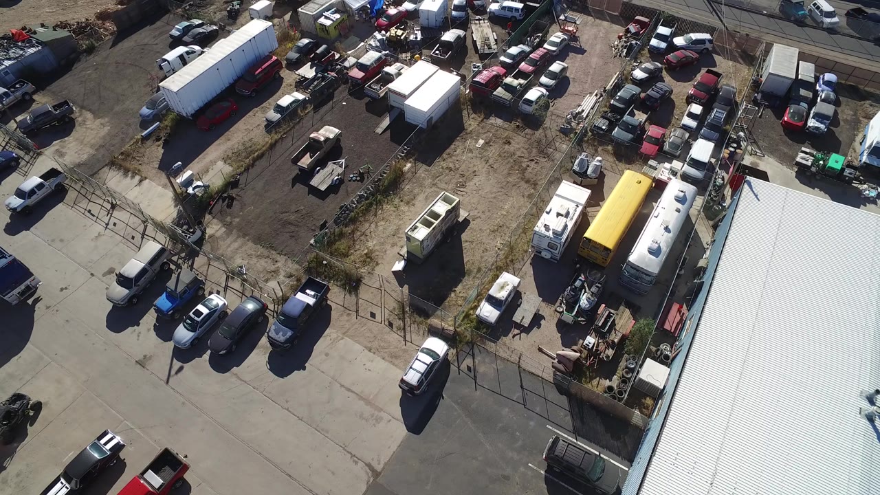 High aerial frame over a Hidden Lake, Colorado auto yard with parked cars, trailers, dirt lots, paved roads, a pale warehouse roof at lower right, and no visible people.