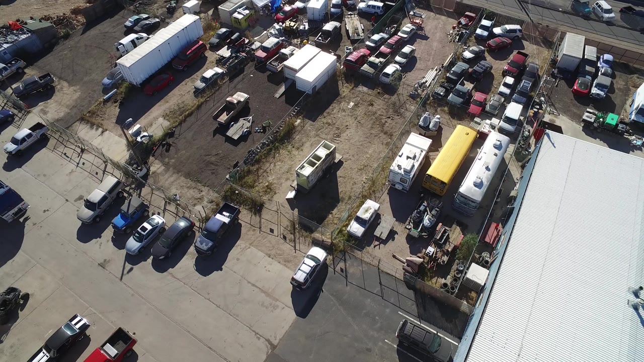 Aerial view in Hidden Lake, Colorado looking down on clustered cars, a yellow trailer, white utility vehicles, fencing, dirt ground, paved lanes, and no visible people.