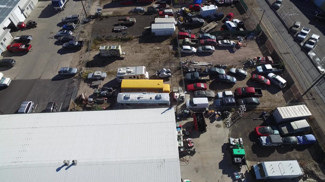 High aerial frame over Hidden Lake, Colorado showing a dirt vehicle lot, rows of parked cars and trucks, a yellow bus-like vehicle, white trailers, a paved road, and no visible people.
