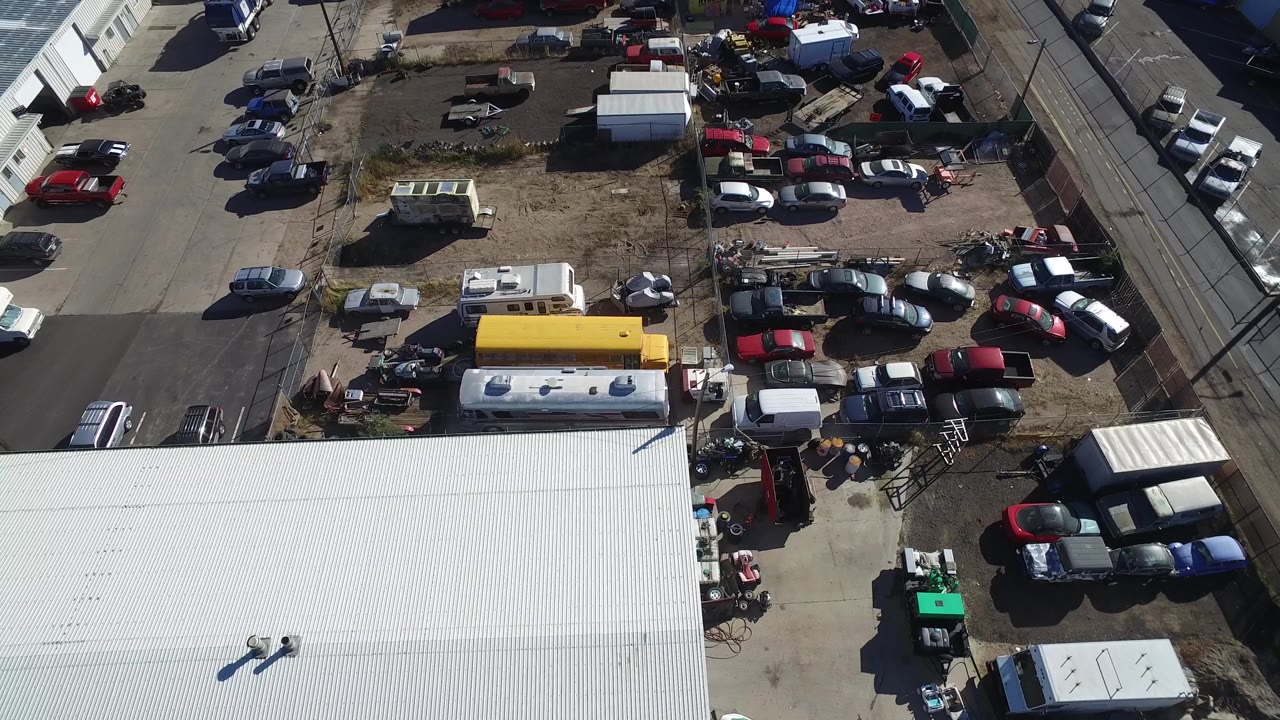 Aerial frame in Hidden Lake, Colorado with parked red, white, black, and gray vehicles clustered around a dirt lot, a yellow bus-like vehicle near the lower center, and no visible people.