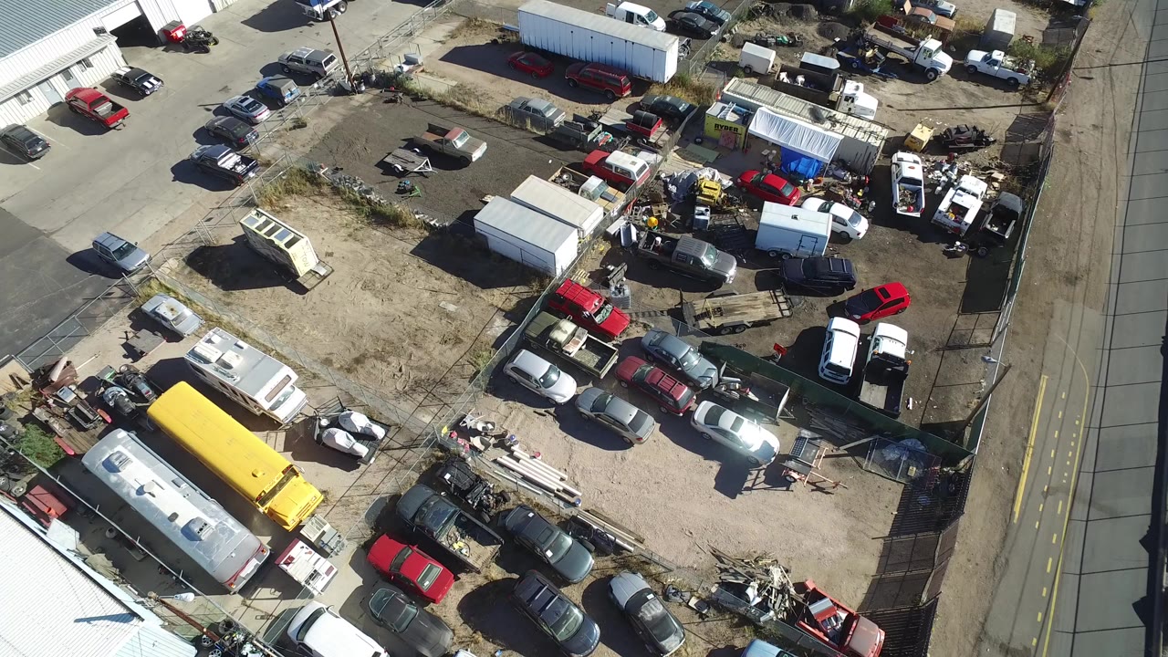 Overhead aerial frame from Hidden Lake, Colorado showing a fenced dirt lot with parked cars, trailers, white sheds, scrap piles, and a paved road along the right edge.