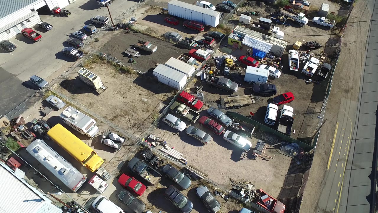 High overhead frame in Hidden Lake, Colorado with the dirt yard centered, white sheds near the middle, a yellow vehicle or trailer at lower left, and rows of parked cars around the lot.