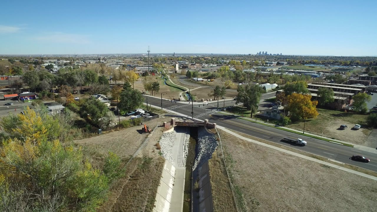 Hidden Lake aerial frame with a multi-lane road crossing the lower view, the drainage channel centered below, small cars on the street, autumn-colored trees, and distant water on the horizon.