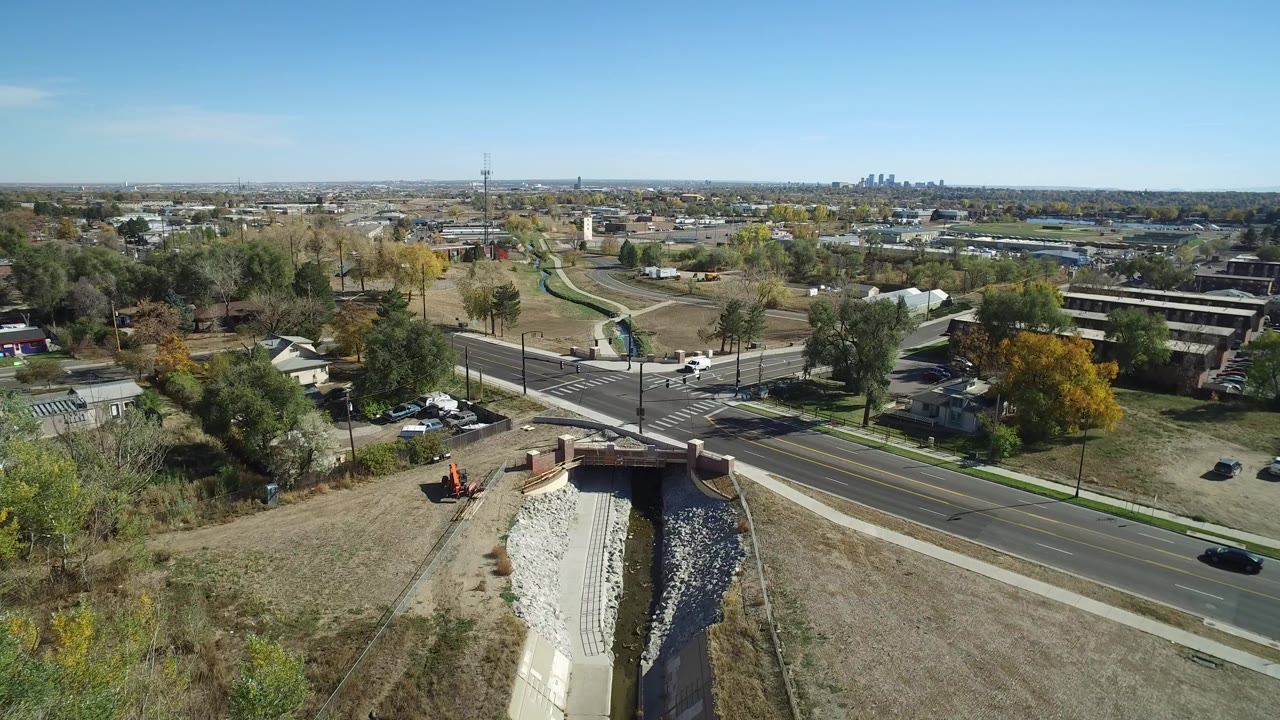 Aerial frame over Hidden Lake, Colorado showing a road, bridge, concrete drainage channel, nearby homes, autumn-toned trees, distant skyline, and no visible people.