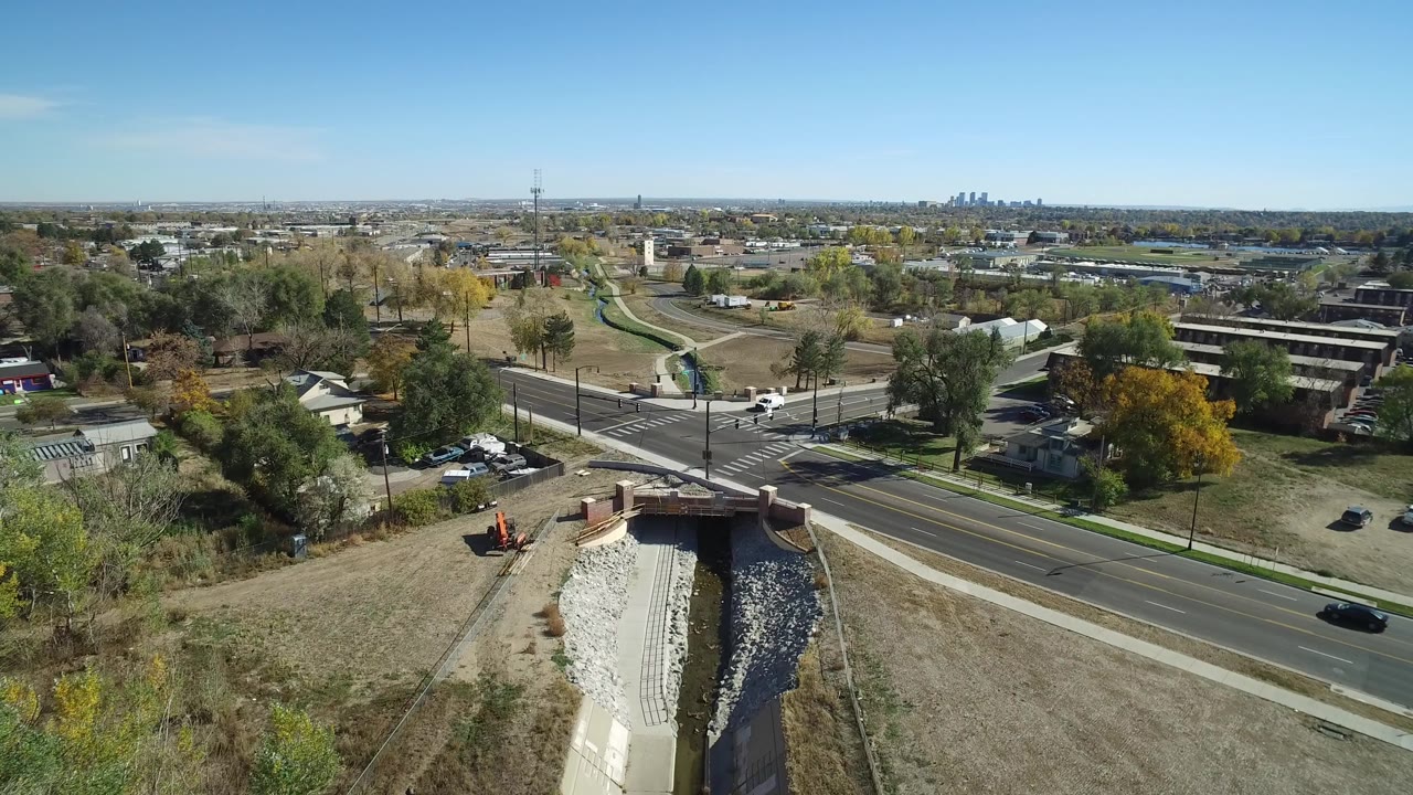Aerial frame in Hidden Lake, Colorado with the drainage channel centered below, a cross street ahead, residential lots, tree cover, a communications tower, distant skyline, and no visible people.