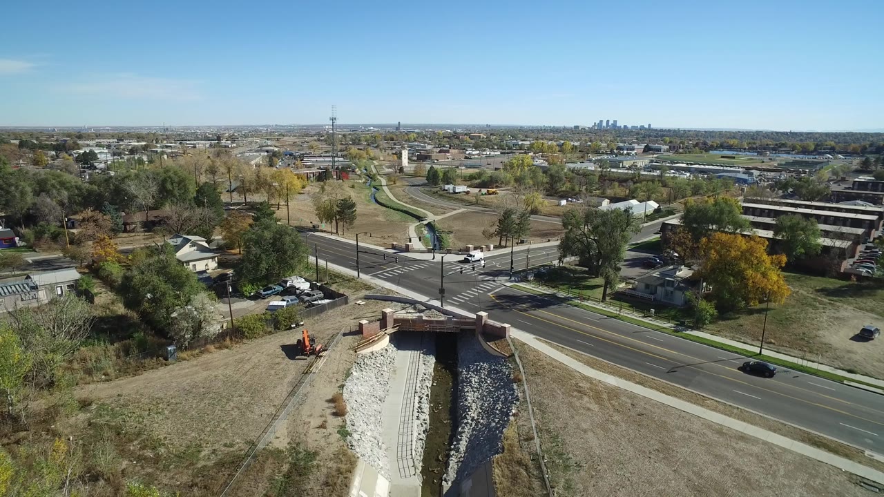 Aerial view over a road bridge, dry grass, homes, trees, and a distant city skyline.