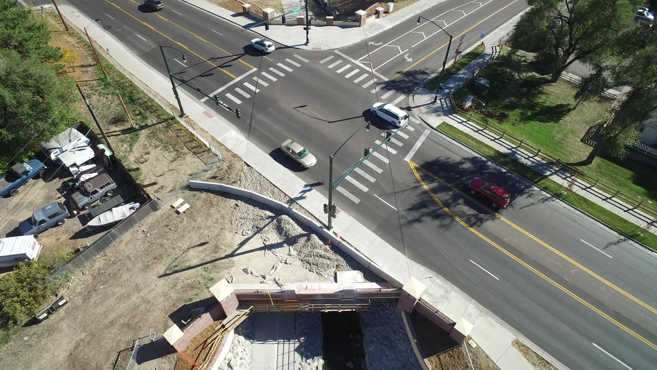 High drone view over a Hidden Lake, Colorado three-way intersection, with a white vehicle near the crosswalk, a red car on the lower road, construction earthwork below, and no visible people.