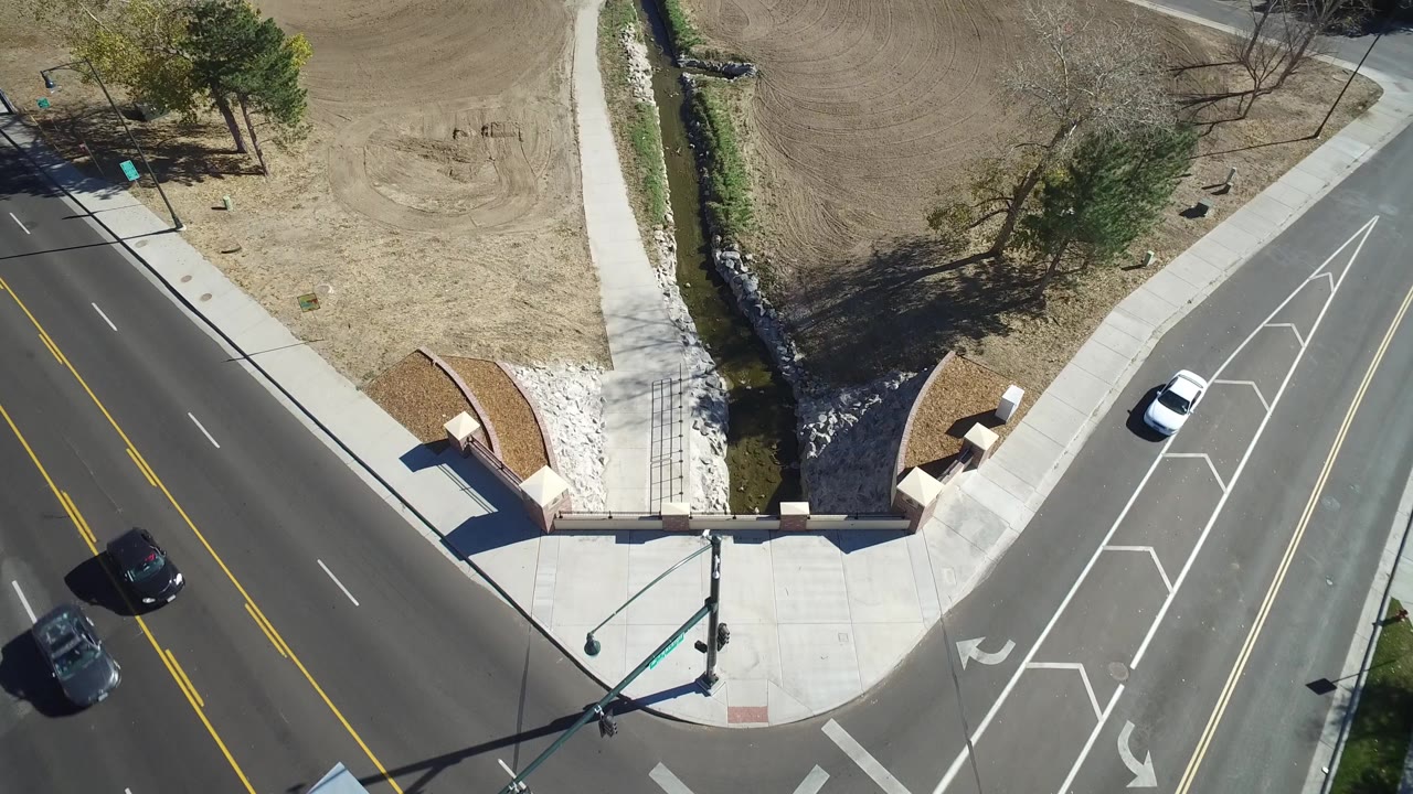 Aerial frame in Hidden Lake, Colorado with the creek and bridge near center, a white car on the road, dry grass, bare soil, trees, and no visible people.
