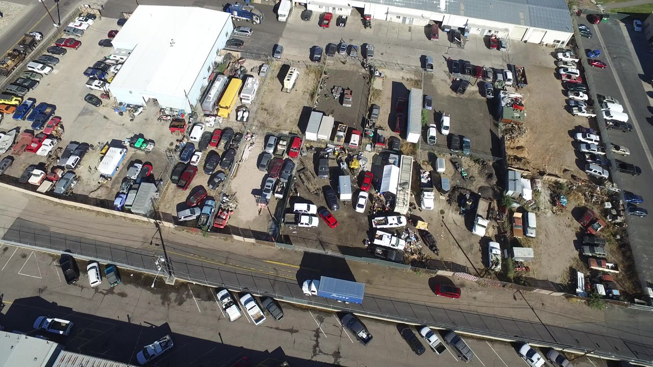 Overhead aerial frame in Hidden Lake, Colorado showing a white-roofed building, packed vehicles, trailers, scrap piles, a bordering road, and no visible people.