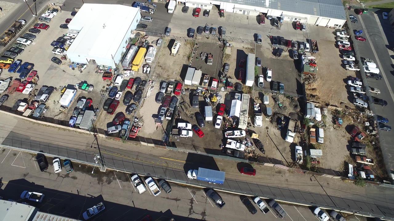 Overhead frame in Hidden Lake, Colorado showing the industrial yard from above, with a road at the bottom, a side street at right, many parked vehicles, and no visible people.