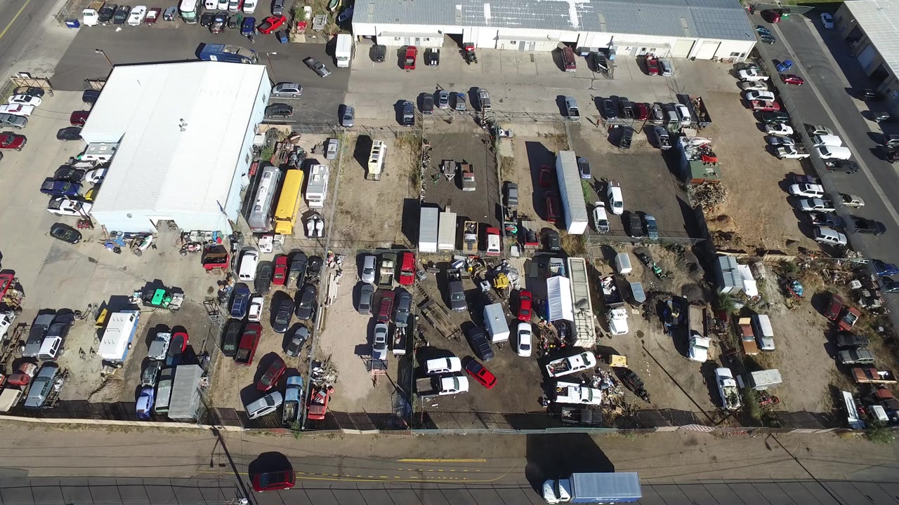 High aerial view in Hidden Lake, Colorado with a large white building on the left, many parked cars, trailers, a yellow vehicle or trailer, road edges, and no visible people.