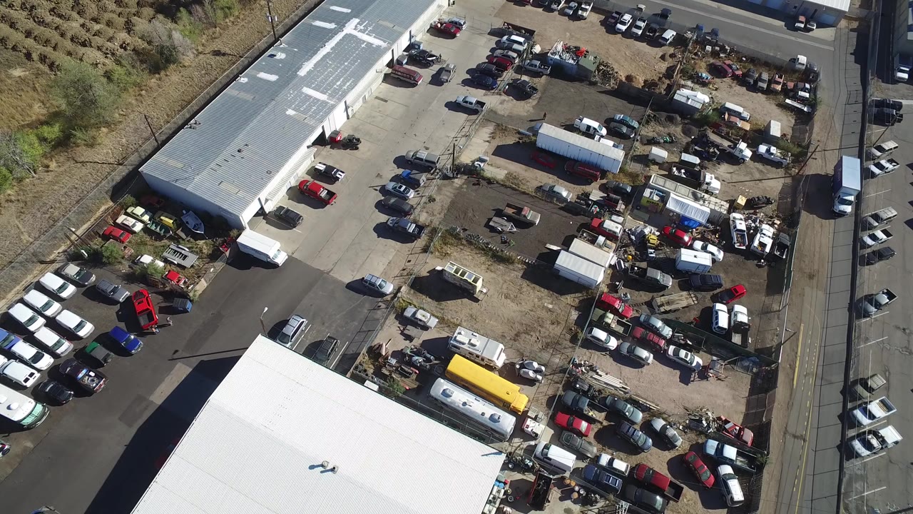 Overhead frame from Hidden Lake, Colorado showing a sunlit industrial parking and storage lot with white-roofed buildings, parked cars and trucks, roadside pavement, and no visible people.