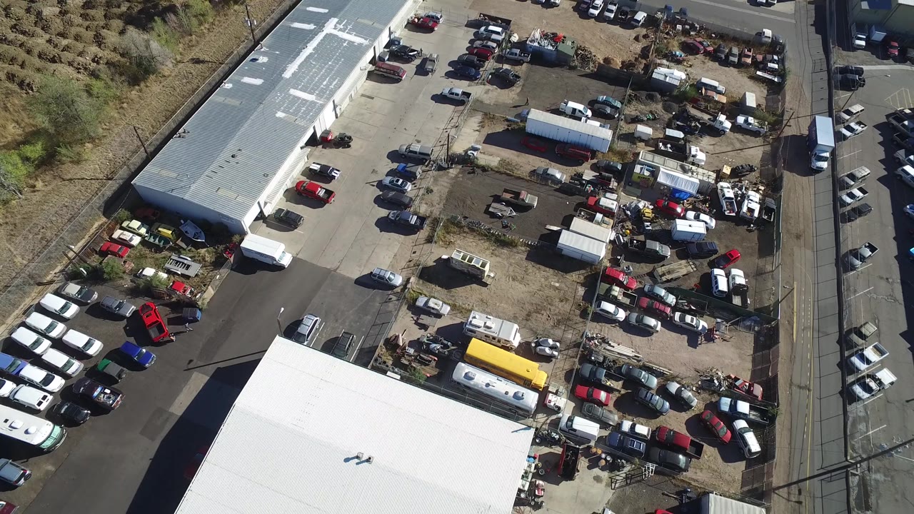 Drone-like overhead view of a vehicle yard beside roads and large roofs.