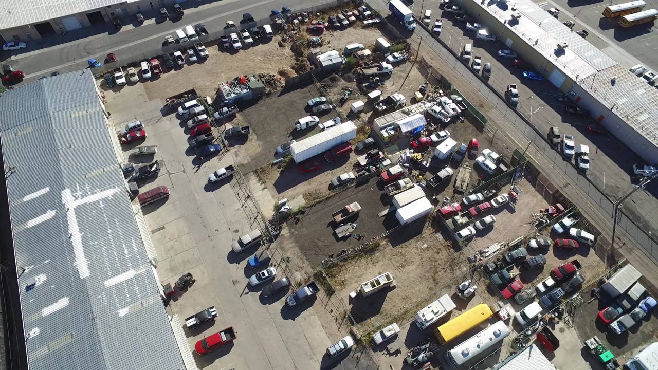 Aerial view in Hidden Lake, Colorado centers on a cluttered commercial yard with rows of parked vehicles, white trailers, warehouse roofs, paved pavement, and no visible people.