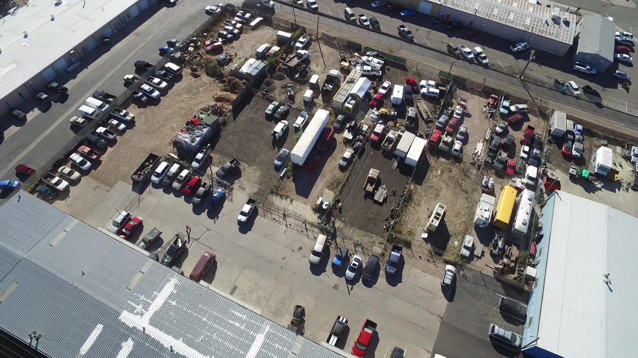 Overhead Hidden Lake, Colorado view with the crowded vehicle lot centered, a pale roof along the bottom edge, and small people visible among cars and equipment.