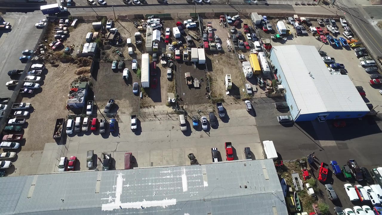 Opening overhead frame of a Hidden Lake, Colorado vehicle yard with parked cars, trailers, equipment piles, a road at the top, and no visible people.