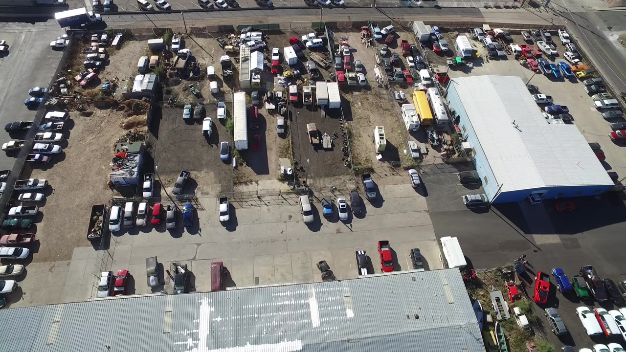 Aerial view of parked cars and work-yard clutter beside two large roofed structures.