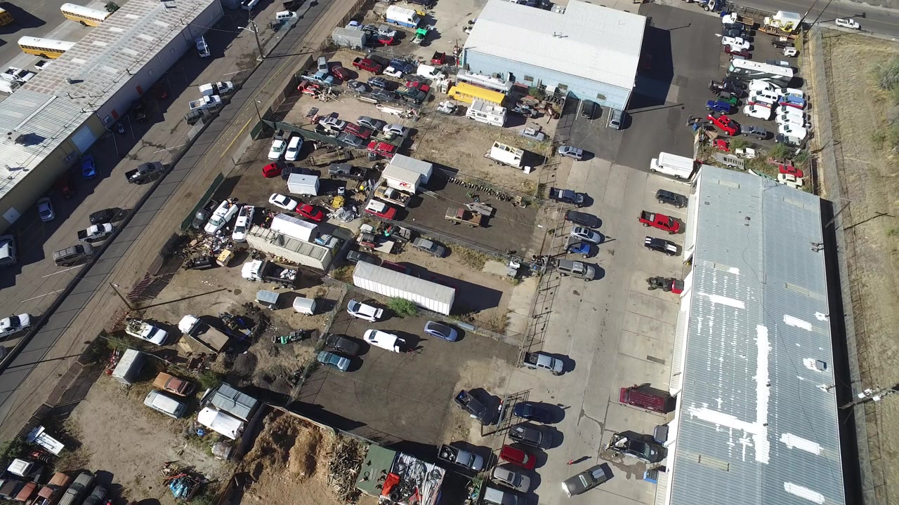 Overhead view of industrial buildings, parked cars, storage piles, and rail tracks.