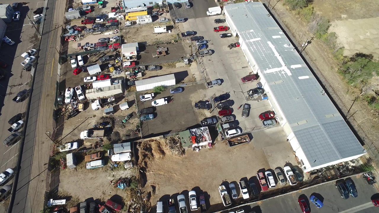 High aerial view in Hidden Lake, Colorado showing a junkyard lot with parked vehicles, scrap piles, small sheds, a long pale-roofed warehouse, road traffic at the left edge, and no visible people.