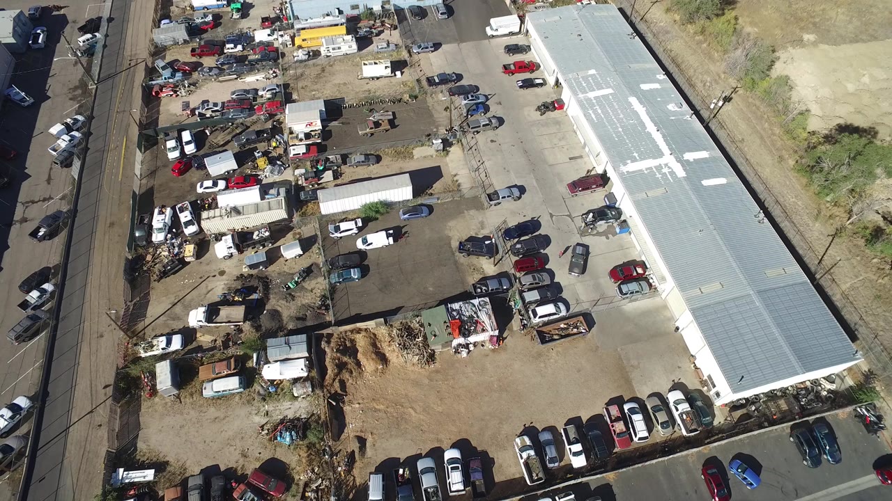 Overhead frame from Hidden Lake, Colorado with rows of parked cars near the bottom, scattered debris and equipment in the center, a long light-gray warehouse on the right, and no visible people.