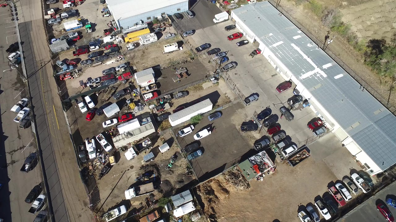 Aerial view of a crowded vehicle lot beside long industrial buildings and a road.