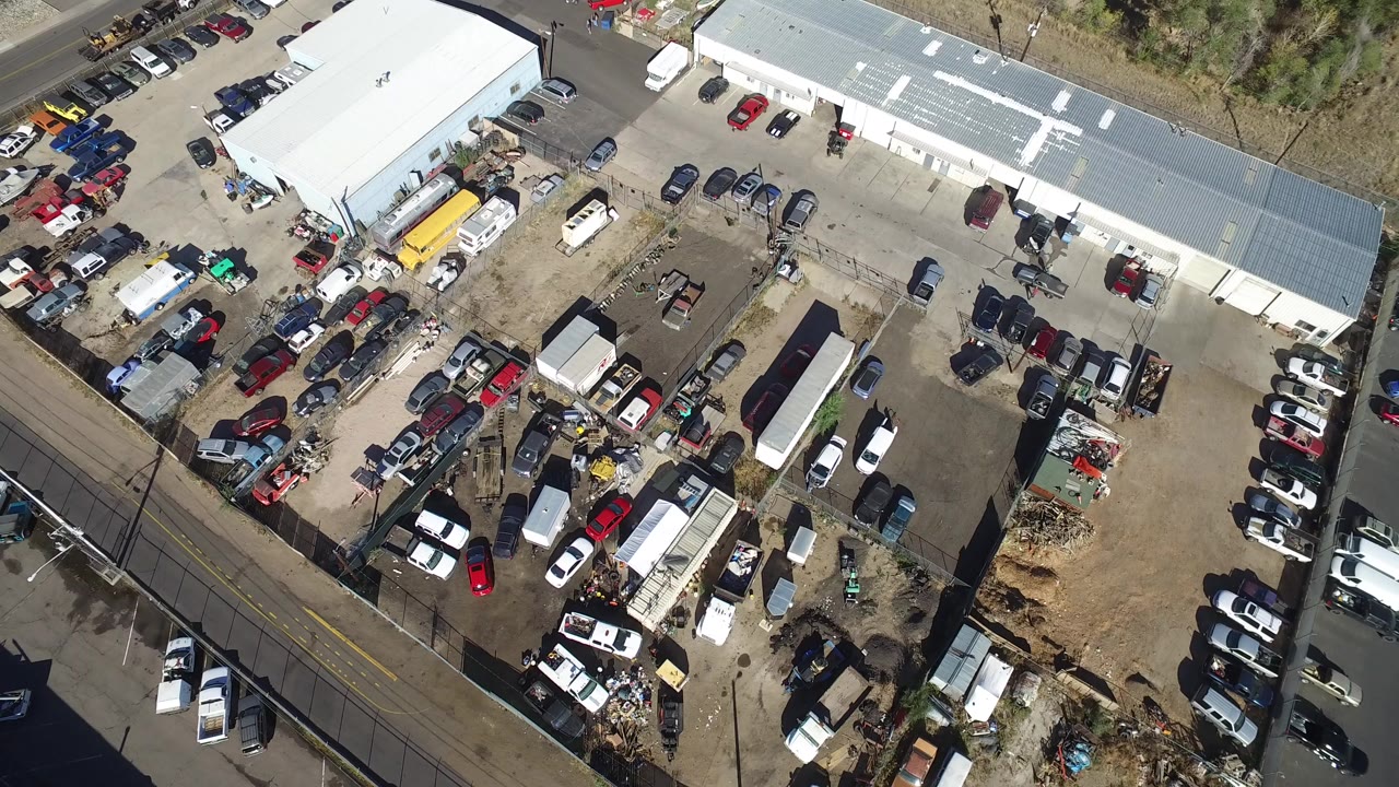 High aerial frame of a Hidden Lake, Colorado workshop yard with white-roofed buildings, parked cars, trailers, boundary walls, adjacent roads, and no visible people.
