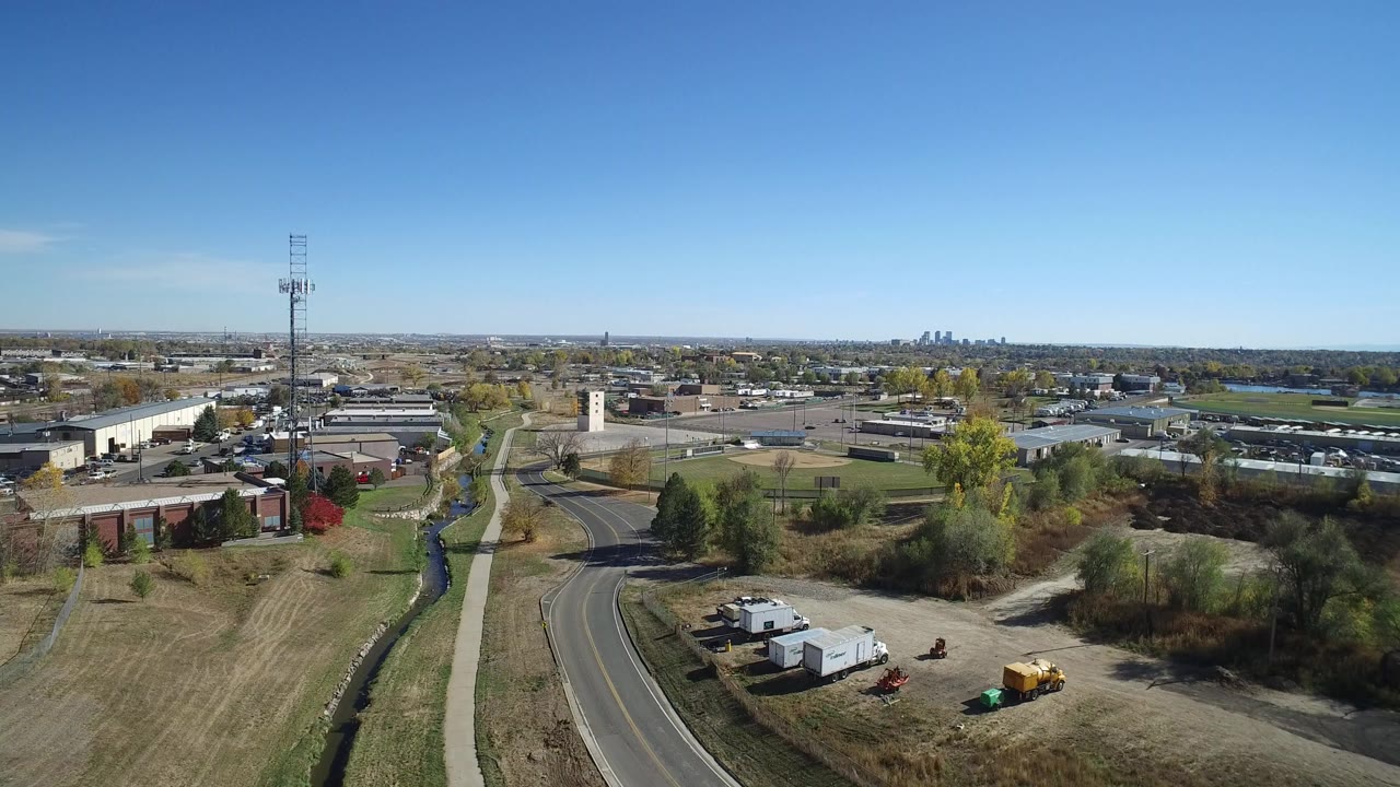 Opening aerial frame over Hidden Lake, Colorado showing a curving road, sidewalk, drainage channel, low buildings, autumn trees, distant skyline, and no visible people.