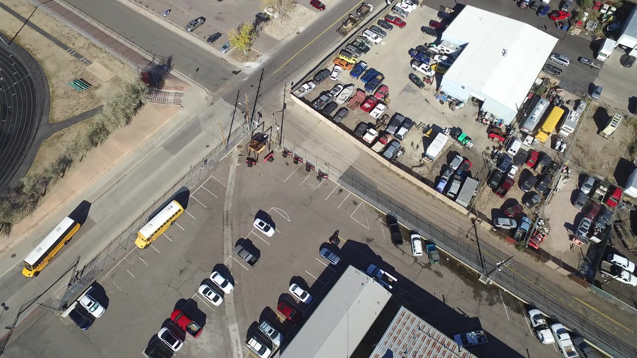 High aerial frame over Hidden Lake, Colorado showing intersecting roads, a parking lot with cars, two yellow school buses, utility poles, and the edge of a track field, with no visible people.