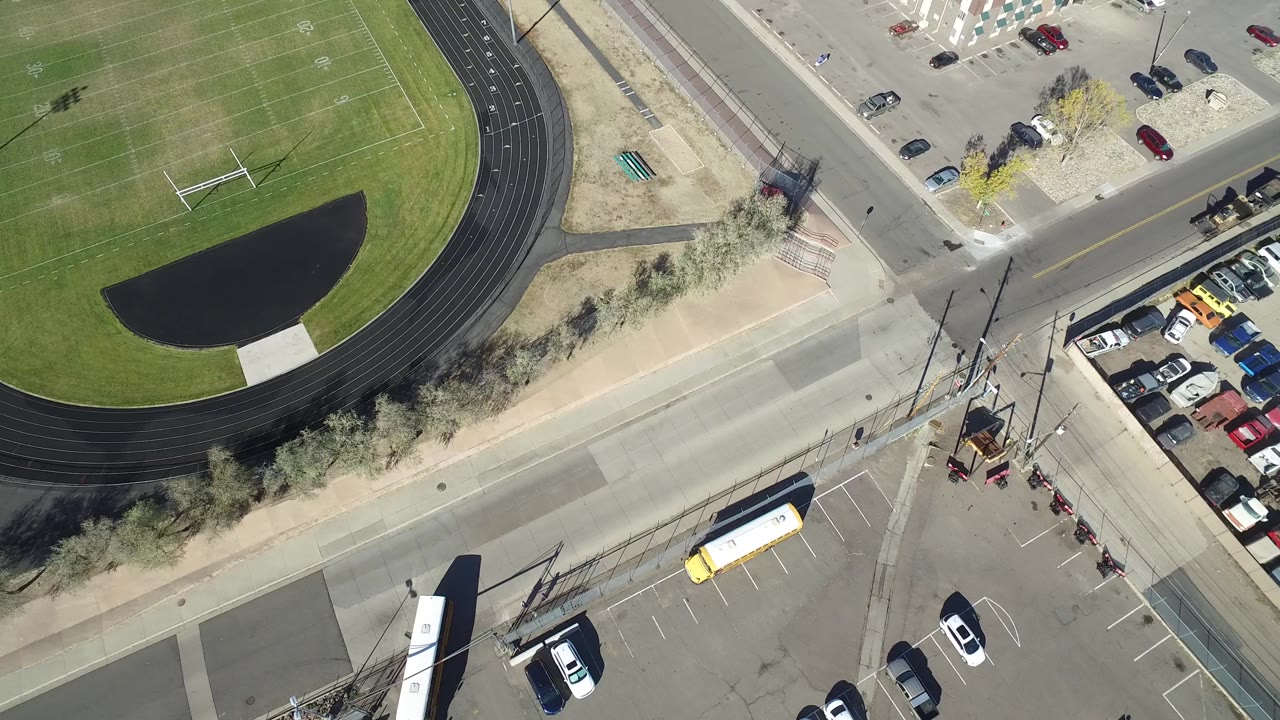 Aerial frame in Hidden Lake, Colorado with the running track and football field prominent, a street crossing below, parked vehicles along the edge, and no visible people.