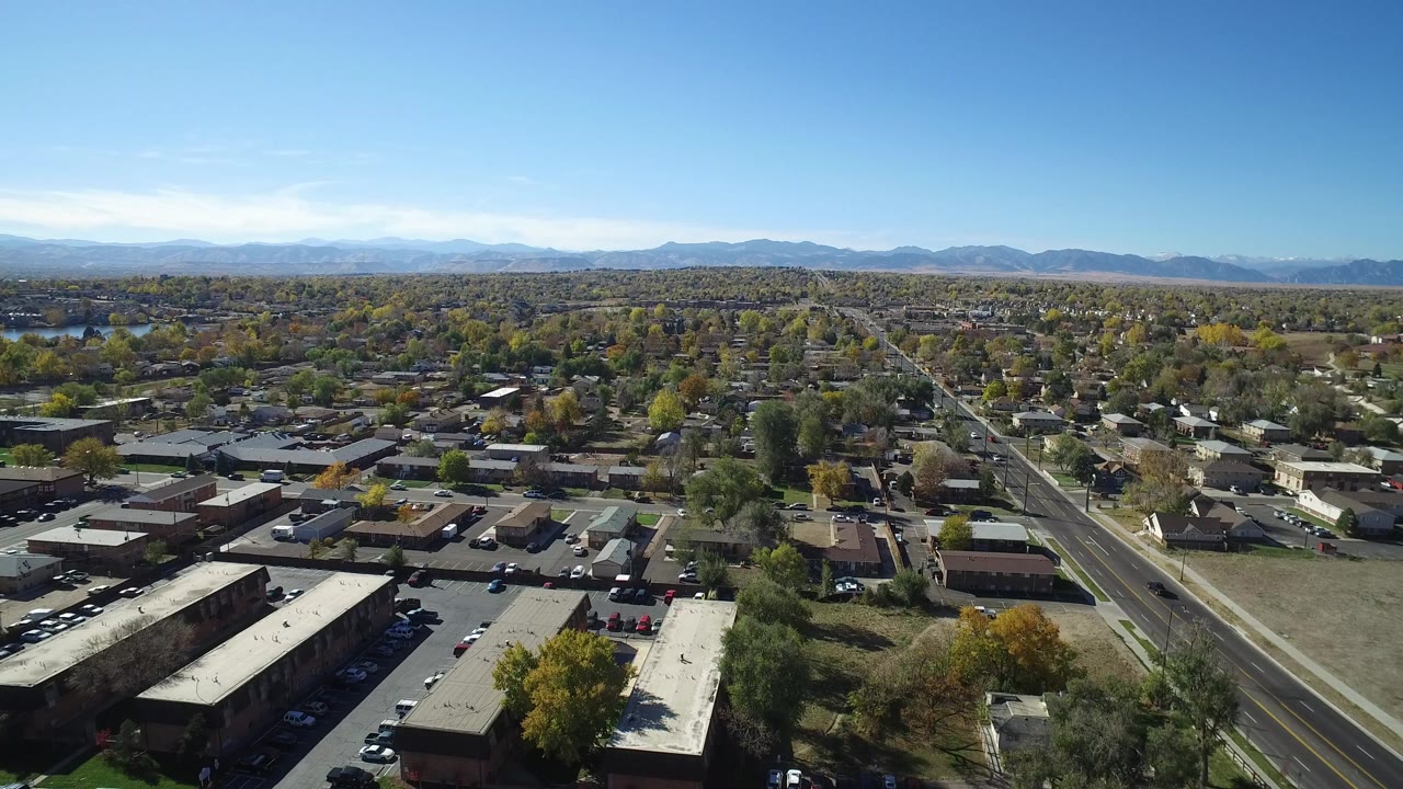 Opening aerial frame over Hidden Lake, Colorado with flat-roof buildings in the foreground, residential streets through the center, distant mountains, blue sky, and no visible people.