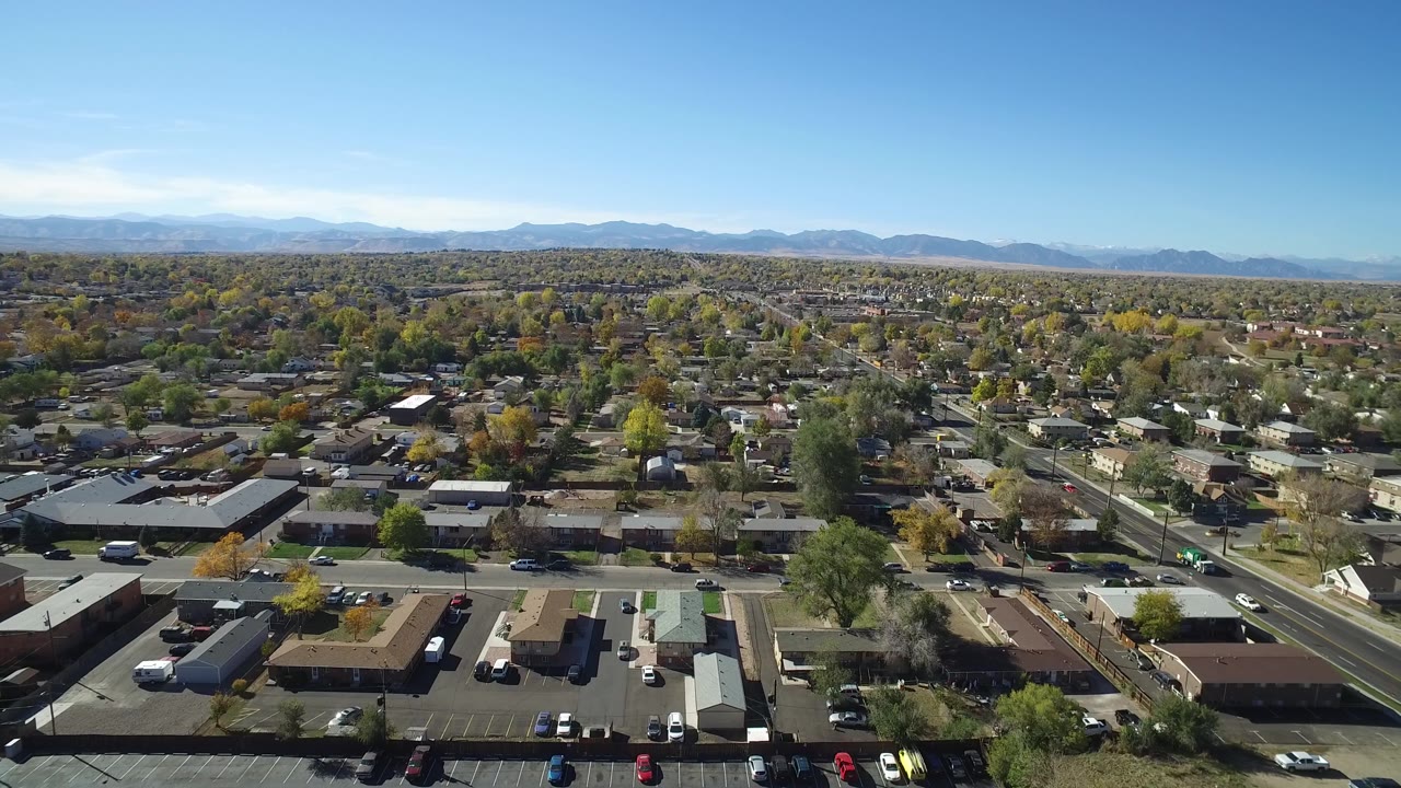 Aerial frame in Hidden Lake, Colorado showing more of the residential grid, scattered autumn trees, low buildings, parked vehicles, a clear sky, and no visible people.