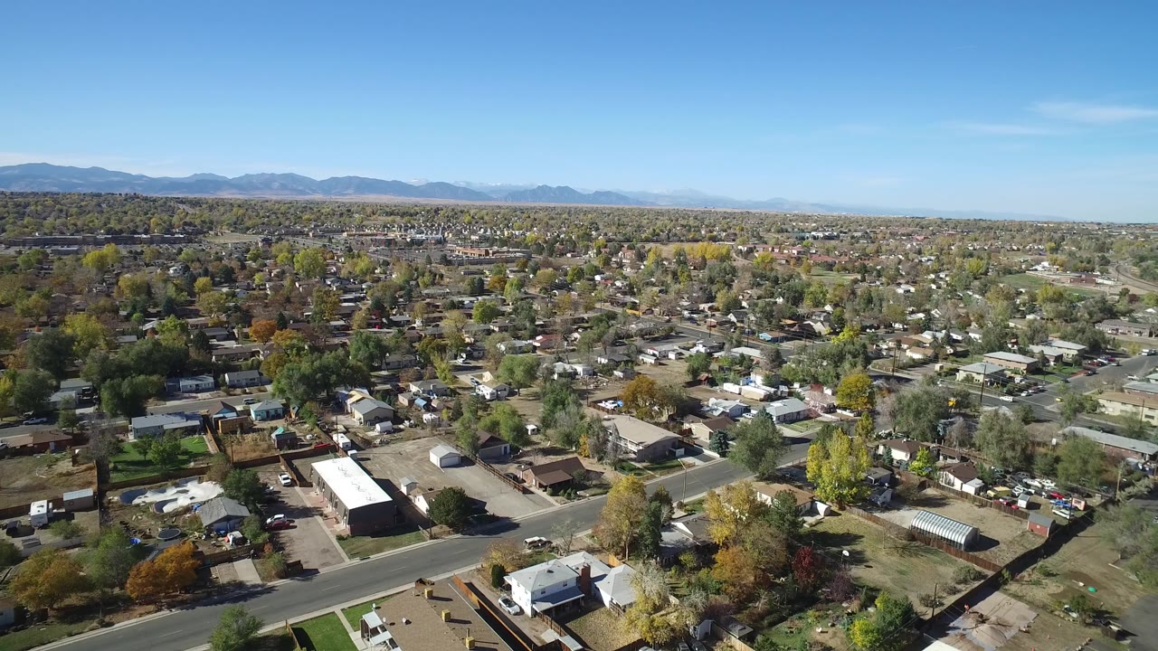 Aerial view in Hidden Lake, Colorado with low residential buildings, a main road in the foreground, scattered cars, autumn-colored trees, mountains, and no visible people.