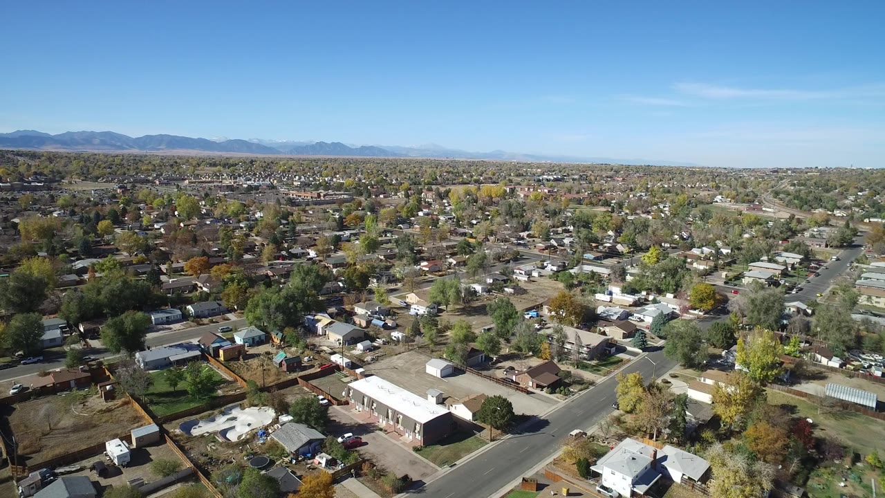 Drone-height frame in Hidden Lake, Colorado showing a road crossing the lower foreground, rows of homes, mixed green and yellow trees, and a mountain line in the distance.