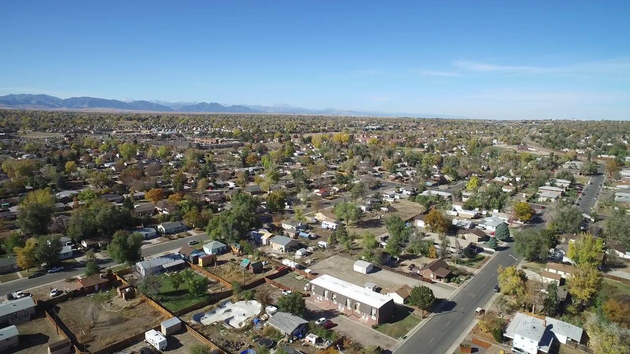 Aerial frame at 0:01 in Hidden Lake, Colorado, showing the residential grid stretching toward a mountain horizon under clear blue sky, with no visible people.
