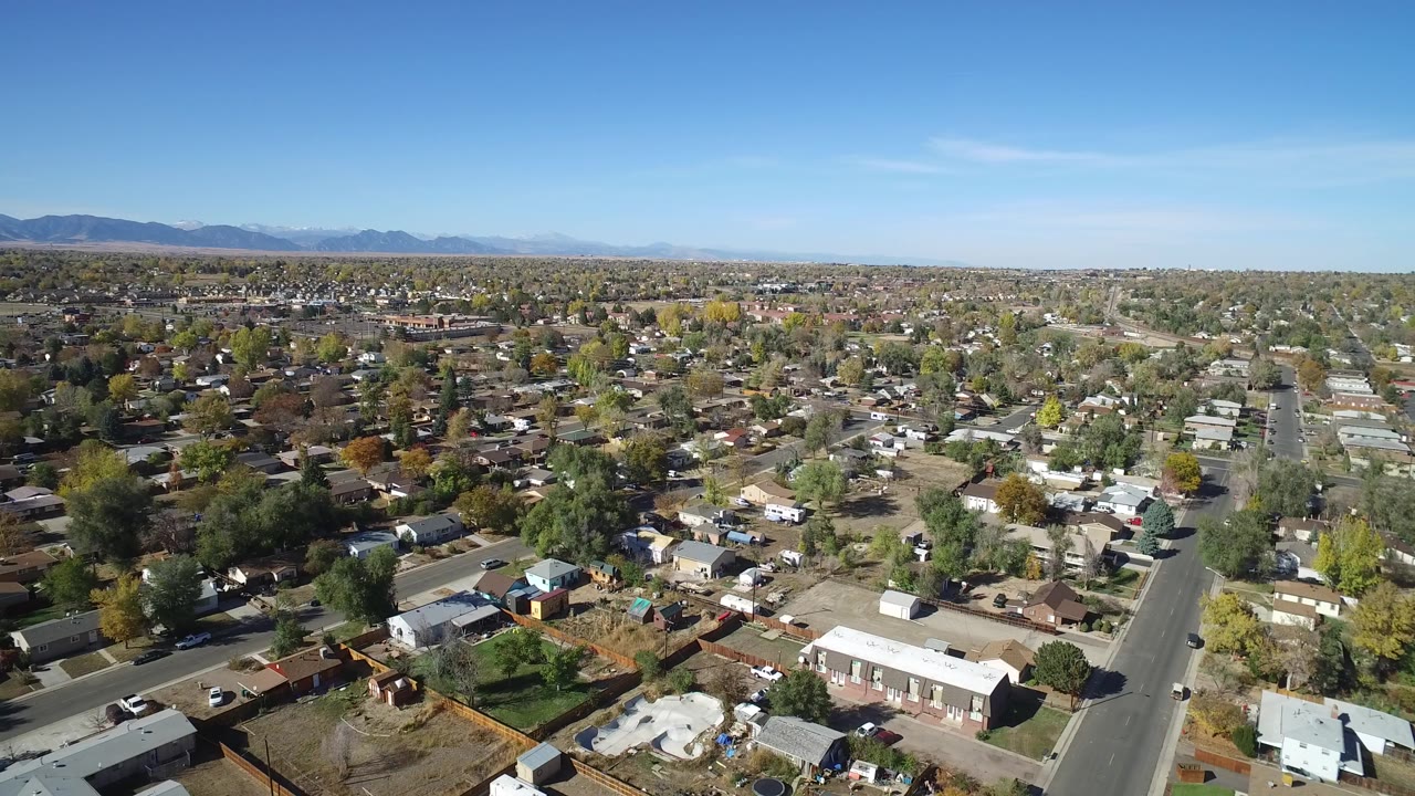 Aerial view over suburban homes, yards, streets, distant mountains, and blue sky.