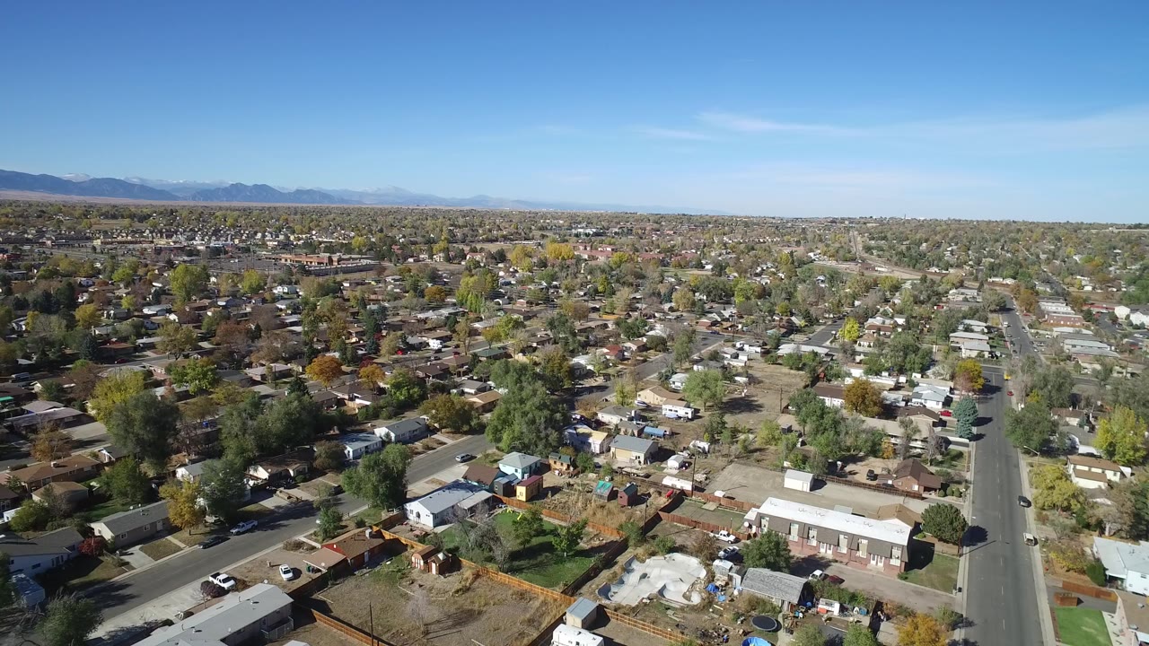 Drone view across Hidden Lake, Colorado with a central paved road, low homes, fenced yards, scattered trees, distant mountains, and no visible people.