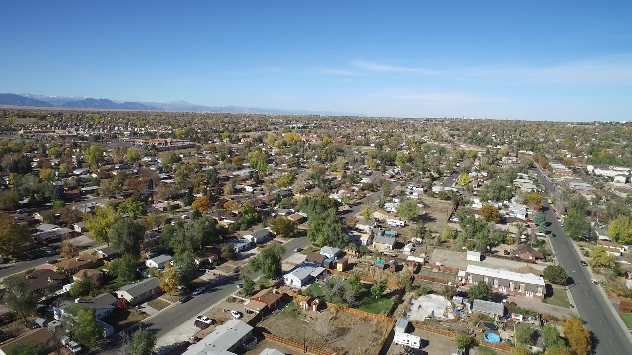 Suburban aerial frame with houses, green and yellow trees, roadways, and open blue sky.