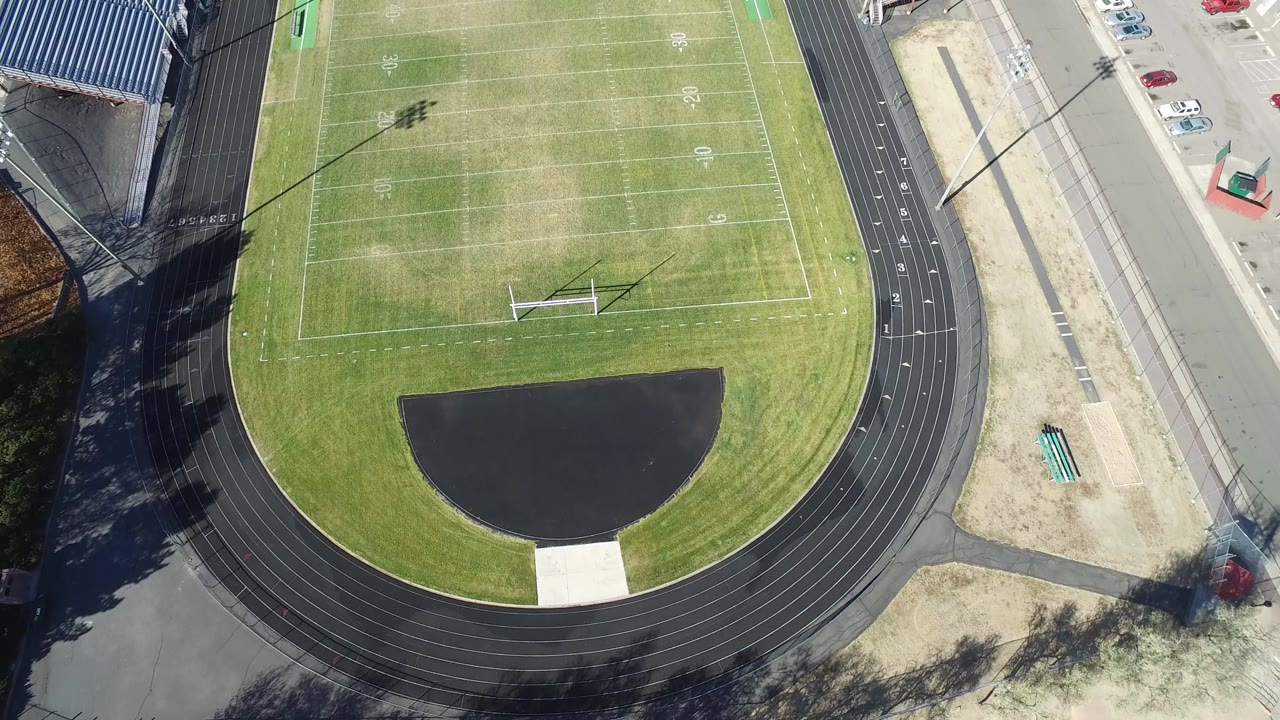 Aerial frame over Hidden Lake, Colorado with the football field centered, the black track curving along the bottom, bleachers at the upper right, and no visible people on the turf or pavement.