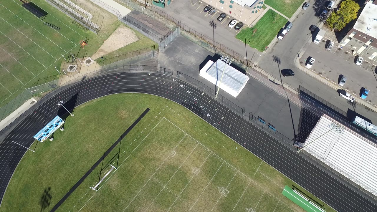 High overhead frame of a Hidden Lake, Colorado sports complex with a black running track, marked football field, fenced courts, bleachers, roads, parked cars, and no visible people.