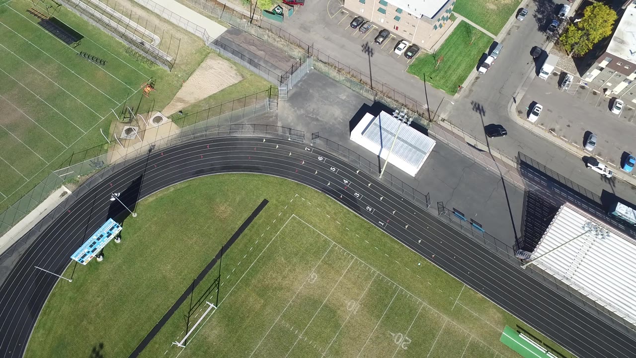Top-down sports field and track with a white-roofed structure, parking area, and fenced courts.