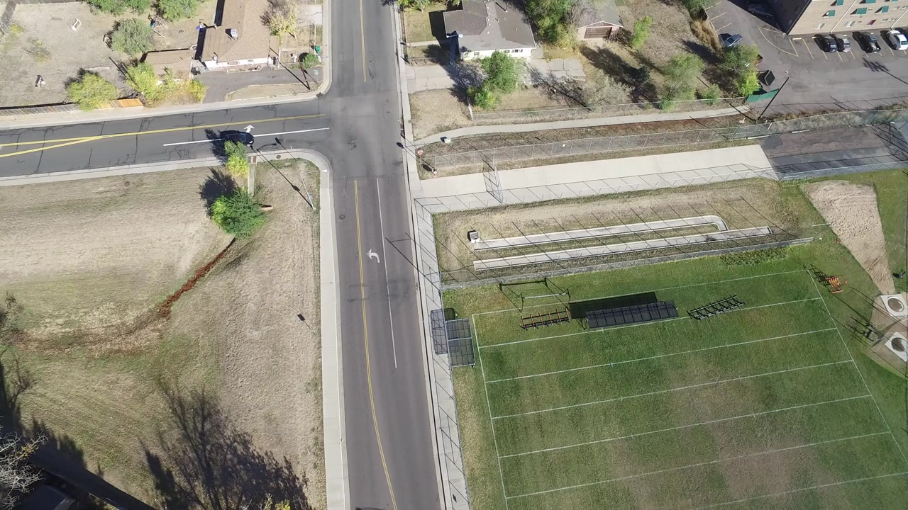 Overhead view in Hidden Lake, Colorado, with a four-way street intersection, marked grass field, sidewalks, dry lawn, trees, nearby buildings, and no visible people.