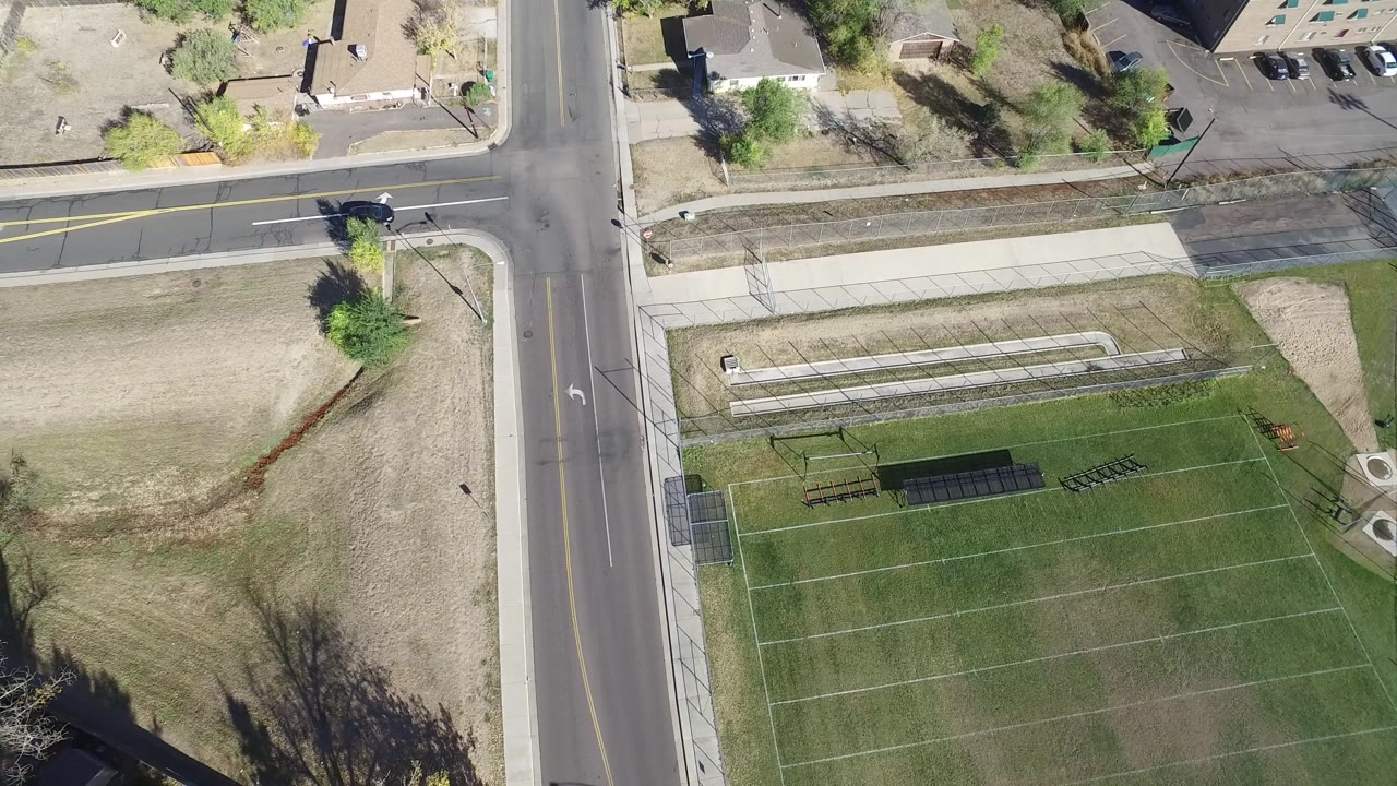 Aerial frame in Hidden Lake, Colorado, showing the road crossing shifted slightly across the view, with the sports field below, bleachers or benches near the field, and no visible people.
