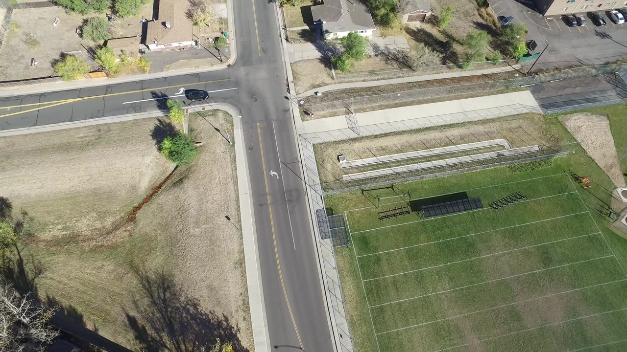 High-angle frame in Hidden Lake, Colorado, with the intersection near the left side, a long sidewalk band above the field, dry open grass, trees, buildings, and no visible people.