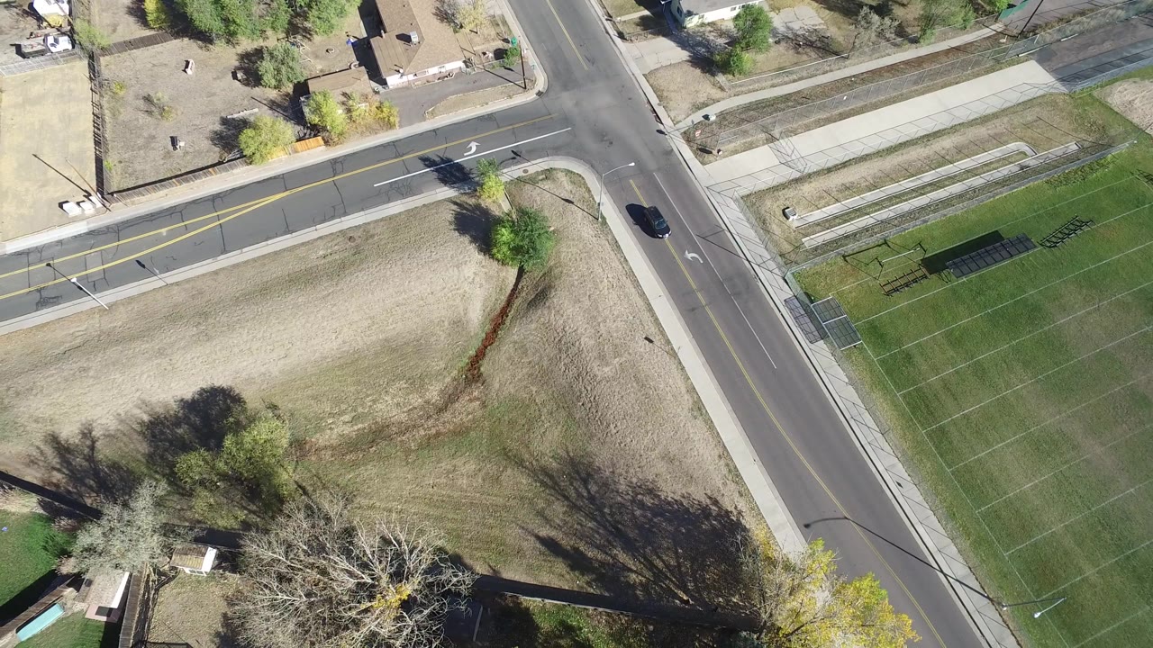 Top-down Hidden Lake, Colorado view showing a dark car farther along the vertical road, a corner sidewalk, sports-field grid lines, dry grass, trees, and no visible people.