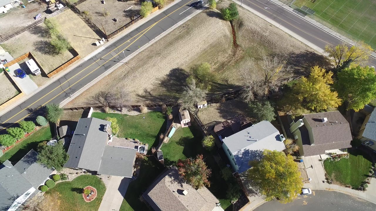 Aerial top-down frame in Hidden Lake, Colorado, with gray and brown rooftops, a landscaped lawn, parked vehicles, roadside trees, and no visible people.
