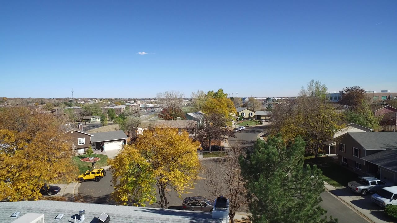 Aerial view over homes, autumn trees, streets, and a yellow vehicle under a clear sky.