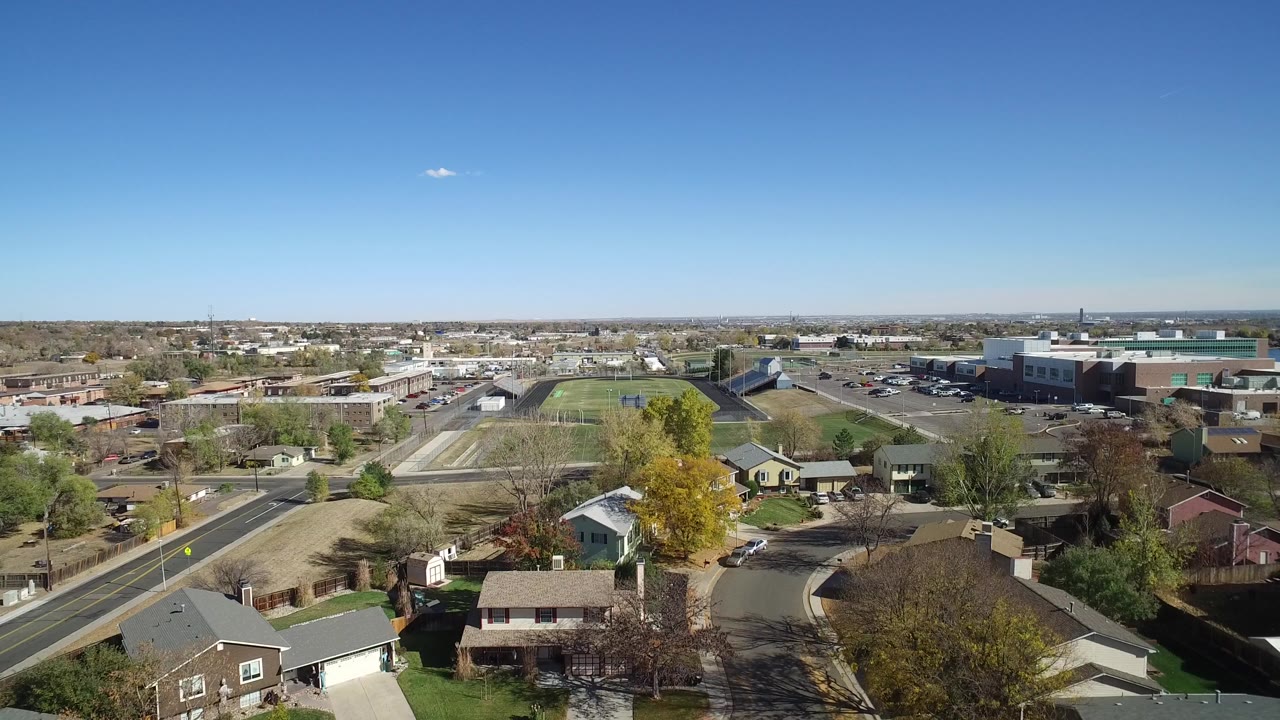 Aerial view over suburban houses, roads, sports fields, and low buildings under a blue sky.