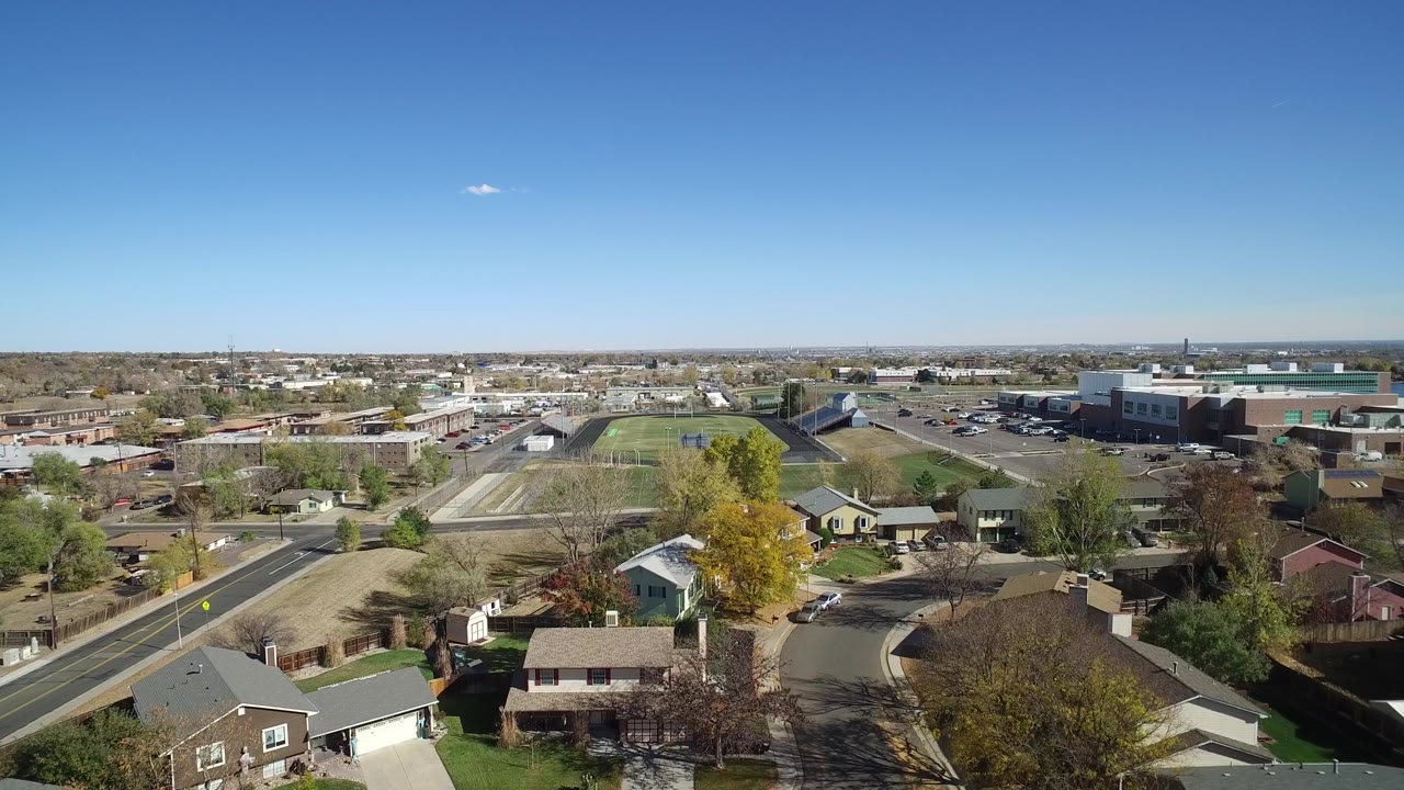 Aerial frame over Hidden Lake, Colorado, with rooftops and trees in the lower frame, sports fields near center, parking lots to the right, and no visible people.