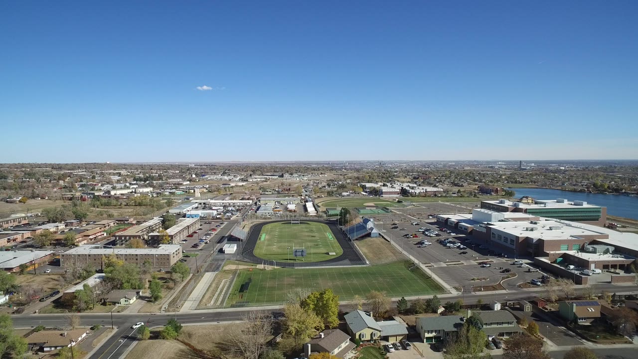 Opening aerial frame over Hidden Lake, Colorado, with the football field centered near low buildings, parking lots, homes, a lake at right, and no visible people.